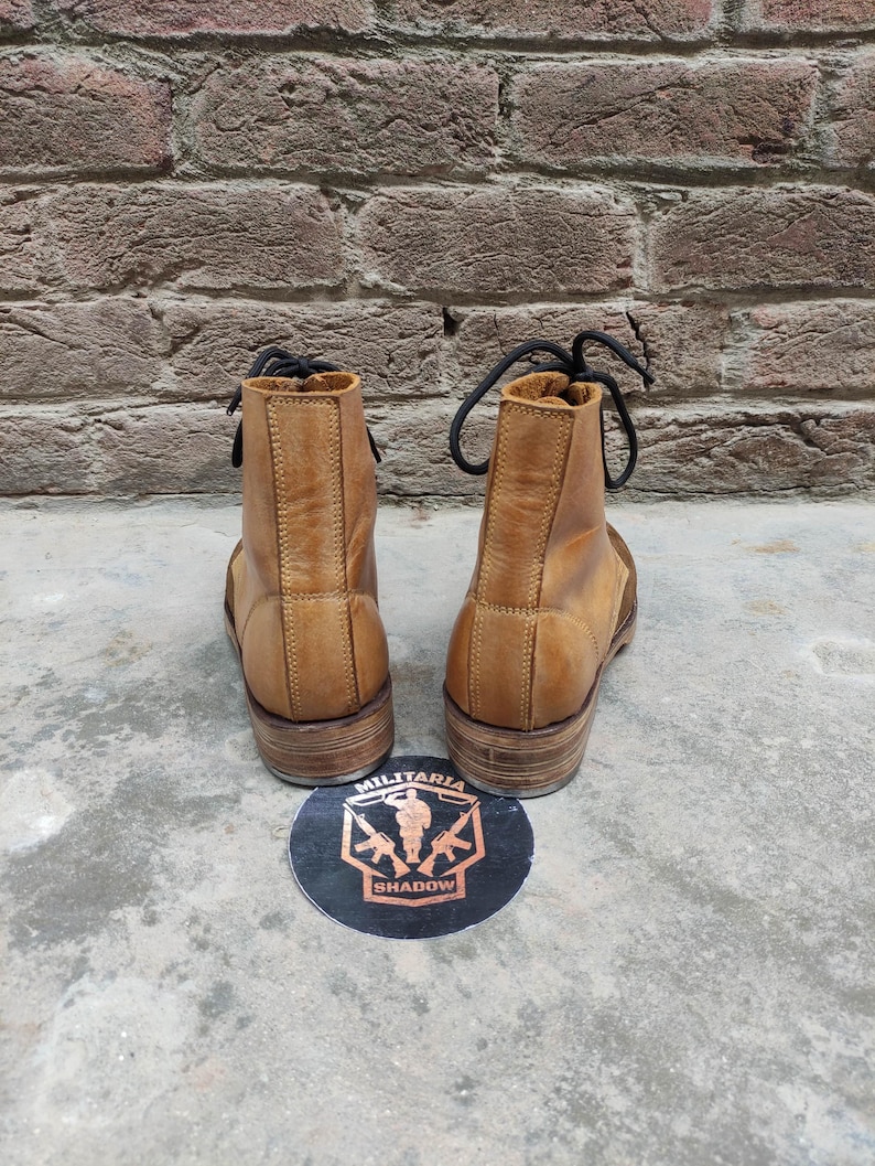 May include: A pair of tan leather boots with black laces, viewed from the back. The boots have a worn appearance and are set against a brick wall background. A round sticker with the words "MILITARIA SHADOW" is on the ground.