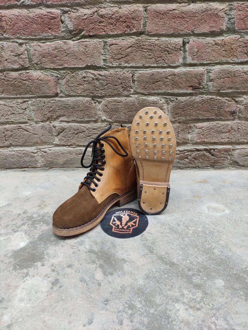 May include: A pair of brown and tan leather boots with black laces. One boot is upright, and the other is lying on its side, revealing the studded sole. A circular logo with text is visible.