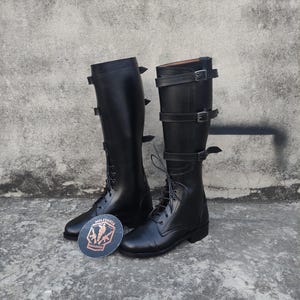 May include: Two black leather riding boots, featuring lace-up fronts and buckle straps. The knee-high boots have a traditional design. A round sticker with "MILITARIA SHADOW" is on the ground.