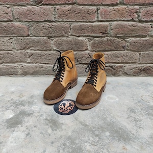 May include: A pair of two-tone leather boots with brown suede vamps and tan uppers, laced with black laces. The boots are set on a concrete surface with a brick wall background. A circular patch with text and a logo is visible.