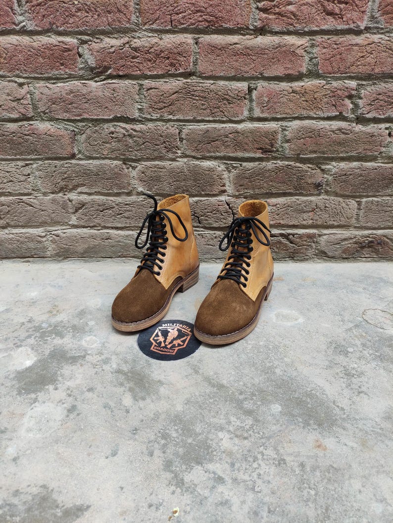 May include: A pair of two-tone leather boots with brown suede vamps and tan uppers, laced with black laces. The boots are set on a concrete surface with a brick wall background. A circular patch with text and a logo is visible.
