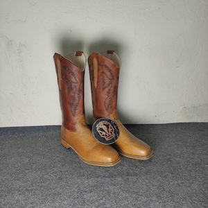 Puede incluir: Un par de botas vaqueras de cuero marrón en dos tonos con punta cuadrada y costuras intrincadas en la caña. Una etiqueta negra circular con el texto MILITARIA SHADOW está unida a la parte delantera de la bota derecha, con un gráfico de un soldado.