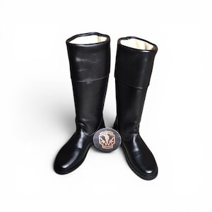 May include: A pair of black leather riding boots with a smooth finish. The boots have a tall shaft and a rounded toe. A round emblem with the word "MILITARIA" is between the boots.