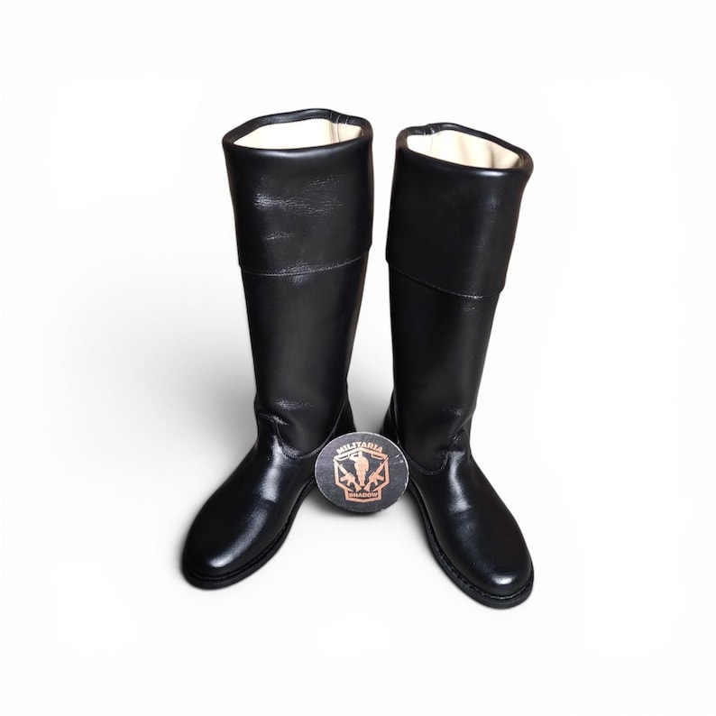 May include: A pair of black leather riding boots with a smooth finish. The boots have a tall shaft and a rounded toe. A round emblem with the word "MILITARIA" is between the boots.