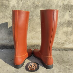 May include: A pair of tall, reddish-brown leather boots. The boots have a smooth finish, a rounded toe, and a flat sole. The boots are standing upright, showcasing their full height and design. A circular emblem with text is visible between the boots.