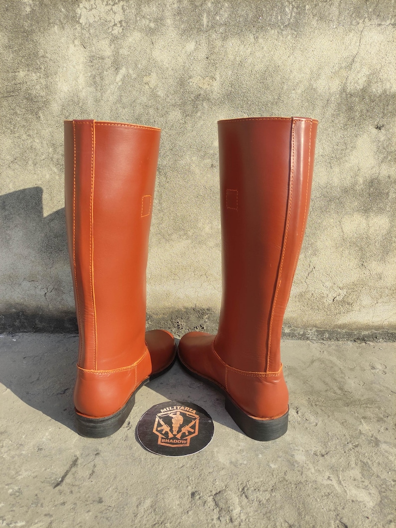 May include: A pair of tall, reddish-brown leather boots. The boots have a smooth finish, a rounded toe, and a flat sole. The boots are standing upright, showcasing their full height and design. A circular emblem with text is visible between the boots.