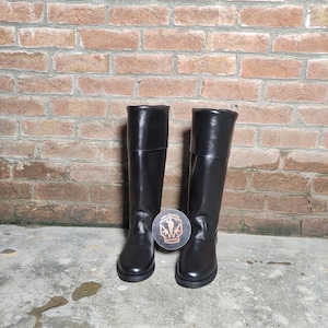 May include: A pair of black leather riding boots. The boots are tall, with a smooth finish and a folded-over top. They are standing on a concrete surface in front of a brick wall. A round coaster with a logo is between the boots.