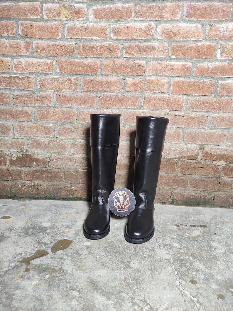 May include: A pair of black leather riding boots. The boots are tall, with a smooth finish and a folded-over top. They are standing on a concrete surface in front of a brick wall. A round coaster with a logo is between the boots.