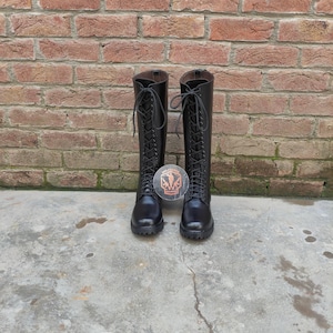 May include: A pair of black, knee-high boots with lace-up closures. The boots are made of a smooth material and have a round toe. A circular sticker with a crest is placed between the boots. The background is a brick wall.