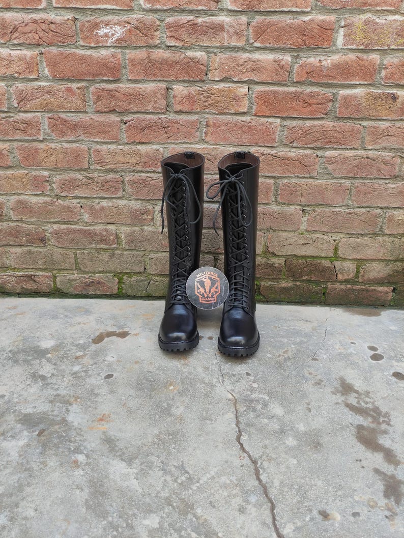 May include: A pair of black, knee-high boots with lace-up closures. The boots are made of a smooth material and have a round toe. A circular sticker with a crest is placed between the boots. The background is a brick wall.