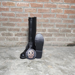 May include: Black leather military-style boots with laces and buckles. The boots are tall, reaching the mid-calf, and are displayed with a round emblem that reads "Militaria Shadow". The boots are set against a brick wall.