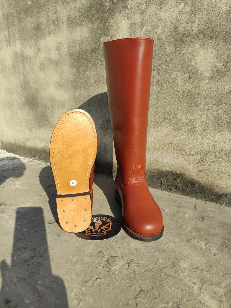 May include: A pair of brown leather riding boots. One boot is upright, the other is lying on its side, revealing the sole. The boots are a reddish-brown color and have a classic design. The sole of the boot is a light tan color. The boots are size 8.