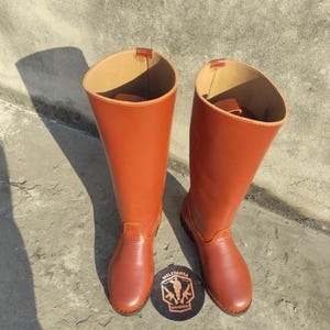 May include: A pair of tall, reddish-brown leather boots. The boots have a smooth finish and a high shaft. A round, black and brown logo with the text "MILITARIA SHADOW" is at the base of the boots.