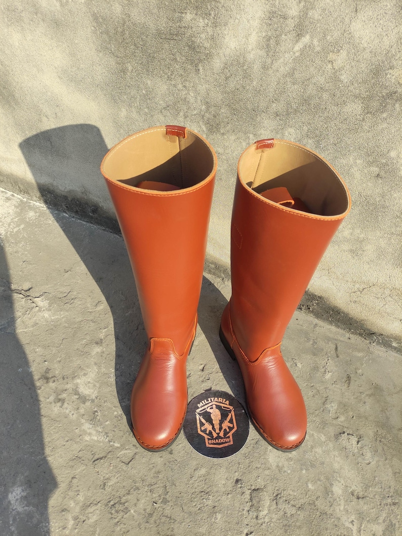 May include: A pair of tall, reddish-brown leather boots. The boots have a smooth finish and a high shaft. A round, black and brown logo with the text "MILITARIA SHADOW" is at the base of the boots.
