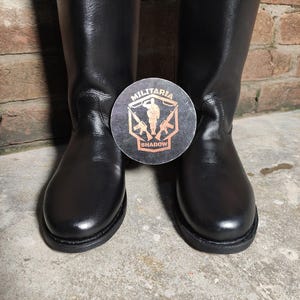May include: A pair of black leather boots with a round toe and a tall shaft. A circular emblem with the text "MILITARIA SHADOW" is placed between the boots. The boots are set against a brick wall and a concrete floor.