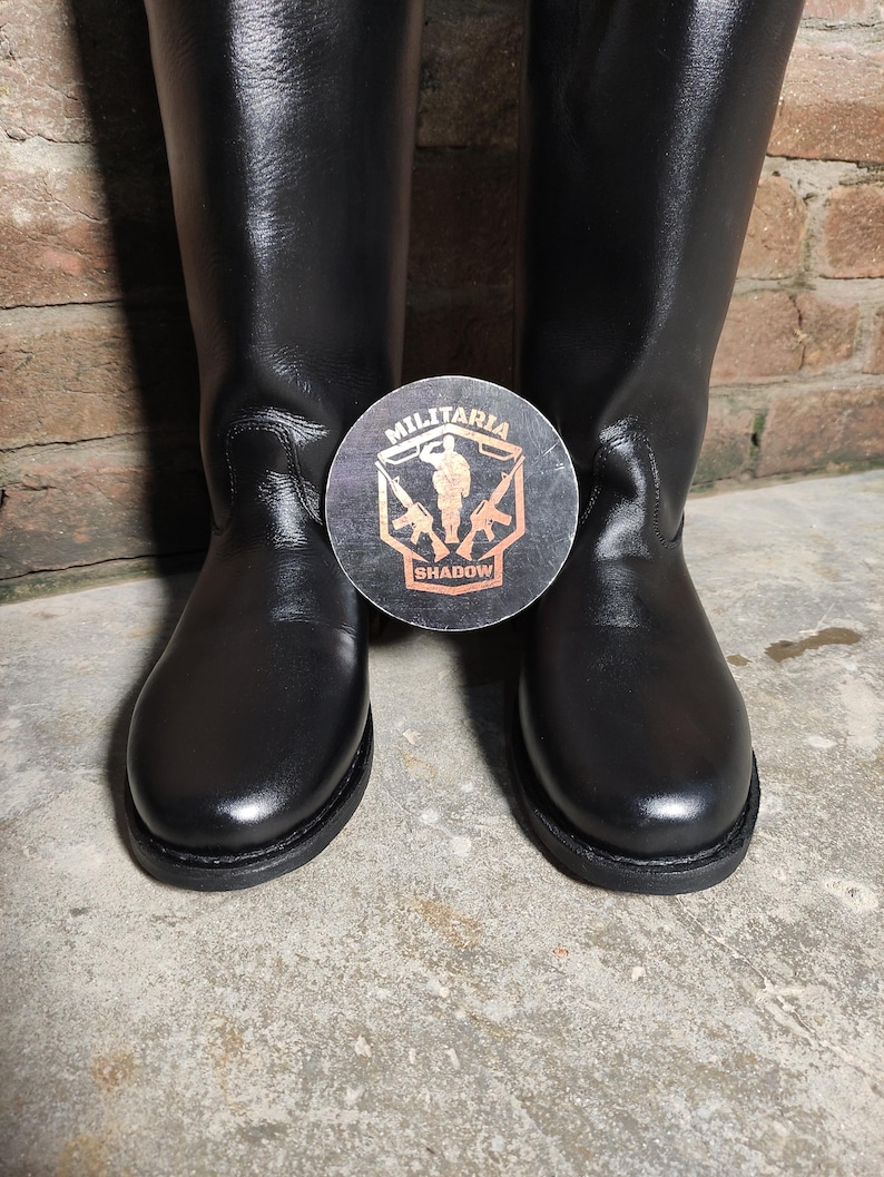 May include: A pair of black leather boots with a round toe and a tall shaft. A circular emblem with the text "MILITARIA SHADOW" is placed between the boots. The boots are set against a brick wall and a concrete floor.