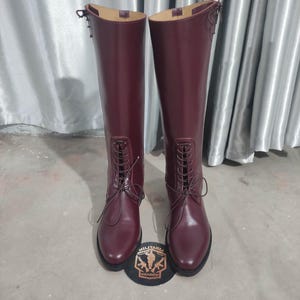 May include: A pair of tall, burgundy leather boots with a lace-up design. The boots have a smooth finish and a rounded toe. A patch with the text "MILITARIA SHADOW" is at the base of the boots.