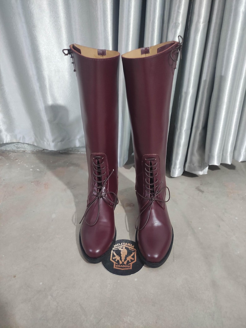 May include: A pair of tall, burgundy leather boots with a lace-up design. The boots have a smooth finish and a rounded toe. A patch with the text "MILITARIA SHADOW" is at the base of the boots.