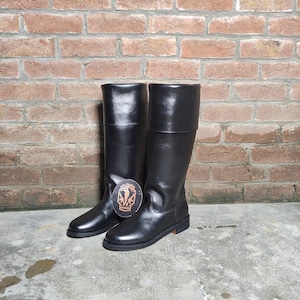 May include: A pair of black leather knee-high boots. The boots have a smooth, shiny finish and a flat sole. The boots are standing on a concrete surface in front of a brick wall.