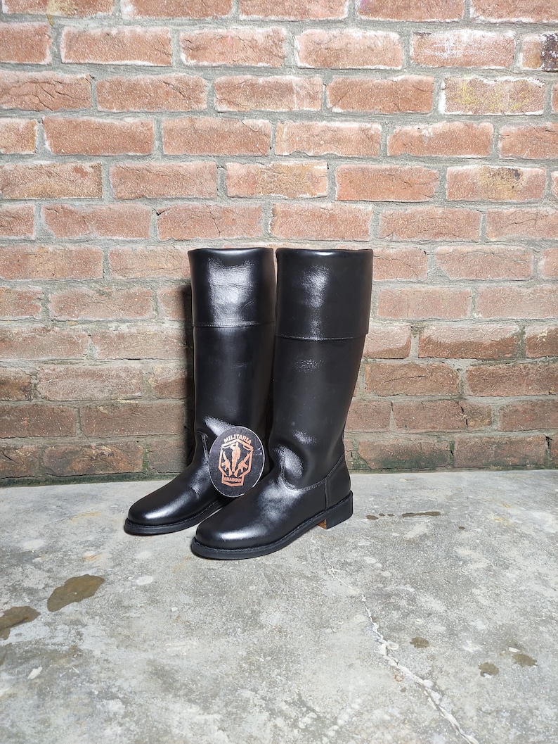 May include: A pair of black leather knee-high boots. The boots have a smooth, shiny finish and a flat sole. The boots are standing on a concrete surface in front of a brick wall.