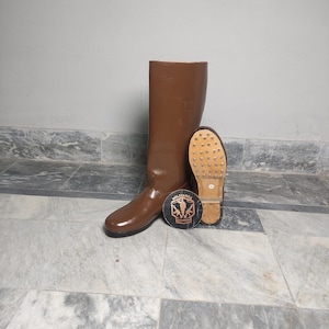 Peut inclure: Une botte en cuir marron avec une semelle assortie et un emblème rond avec le texte "MILITARIA" et un logo. La botte est haute, avec une finition lisse et brillante. La semelle a des clous métalliques.