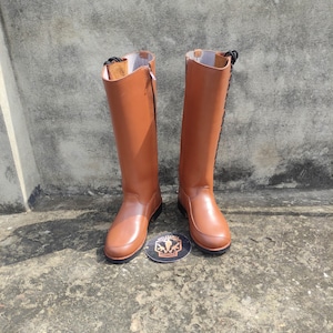 May include: A pair of tall, brown leather boots with a smooth finish. The boots have a zip on one side and laces on the other. The soles are black, and the boots are standing upright. A round emblem is between the boots.