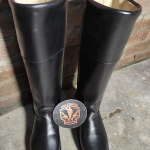 May include: A pair of tall, black leather boots with a smooth finish. The boots have a high shaft and a round toe. The interior lining is a light beige color. A round sticker with the text "MILITARIA SHADOW" is between the boots.