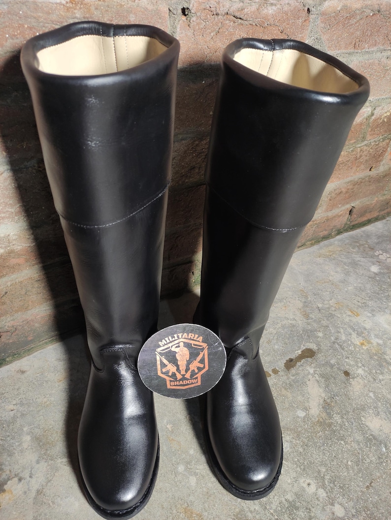 May include: A pair of tall, black leather boots with a smooth finish. The boots have a high shaft and a round toe. The interior lining is a light beige color. A round sticker with the text "MILITARIA SHADOW" is between the boots.