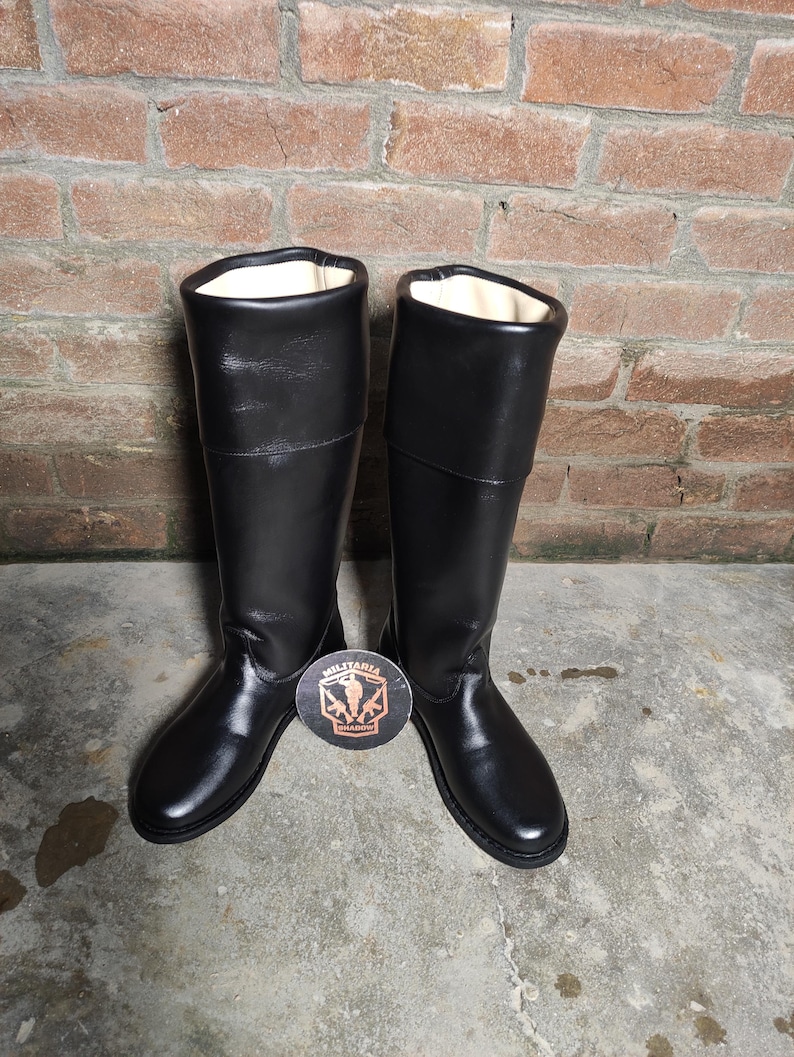 May include: A pair of tall, black leather boots with a smooth, glossy finish. The boots have a high shaft and a rounded toe. The interior lining is a light cream color. A round emblem with text is visible between the boots.