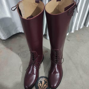 May include: A pair of tall, burgundy leather boots with a lace-up design. The boots have a smooth, glossy finish and are placed on a gray surface. A round emblem with the text "MILITARIA SHADOW" is at the base of the boots.