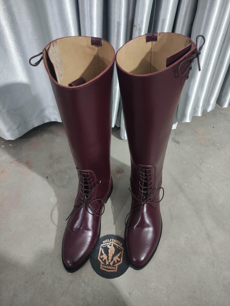 May include: A pair of tall, burgundy leather boots with a lace-up design. The boots have a smooth, glossy finish and are placed on a gray surface. A round emblem with the text "MILITARIA SHADOW" is at the base of the boots.