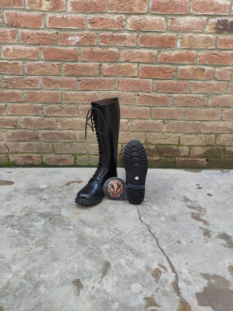 May include: Black leather knee-high boots with a lace-up design. One boot is upright, and the other is lying down, showing the sole. A round emblem with text and a graphic is between the boots. The boots are on a concrete surface in front of a brick wall.