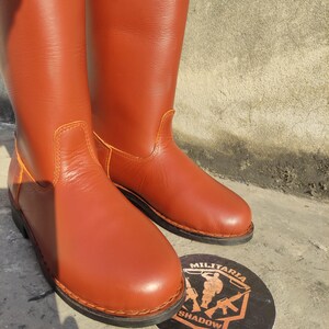 May include: A pair of tall, reddish-brown leather boots. The boots have a smooth finish with visible stitching and a rounded toe. A circular emblem with the text "MILITARIA SHADOW" is visible.