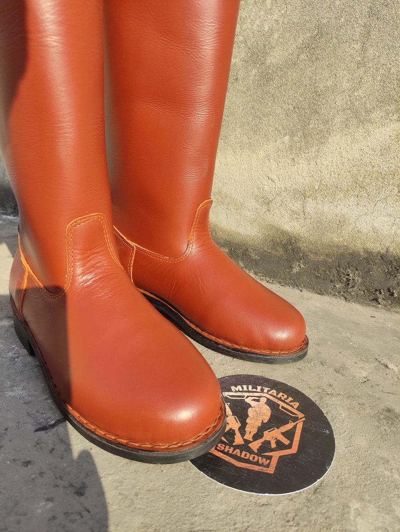 May include: A pair of tall, reddish-brown leather boots. The boots have a smooth finish with visible stitching and a rounded toe. A circular emblem with the text "MILITARIA SHADOW" is visible.