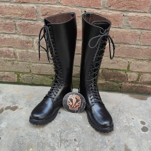 May include: A pair of black leather knee-high boots with lace-up closures. The boots have a rounded toe and a sturdy sole. A round emblem with the text "MILITARIA" is between the boots.