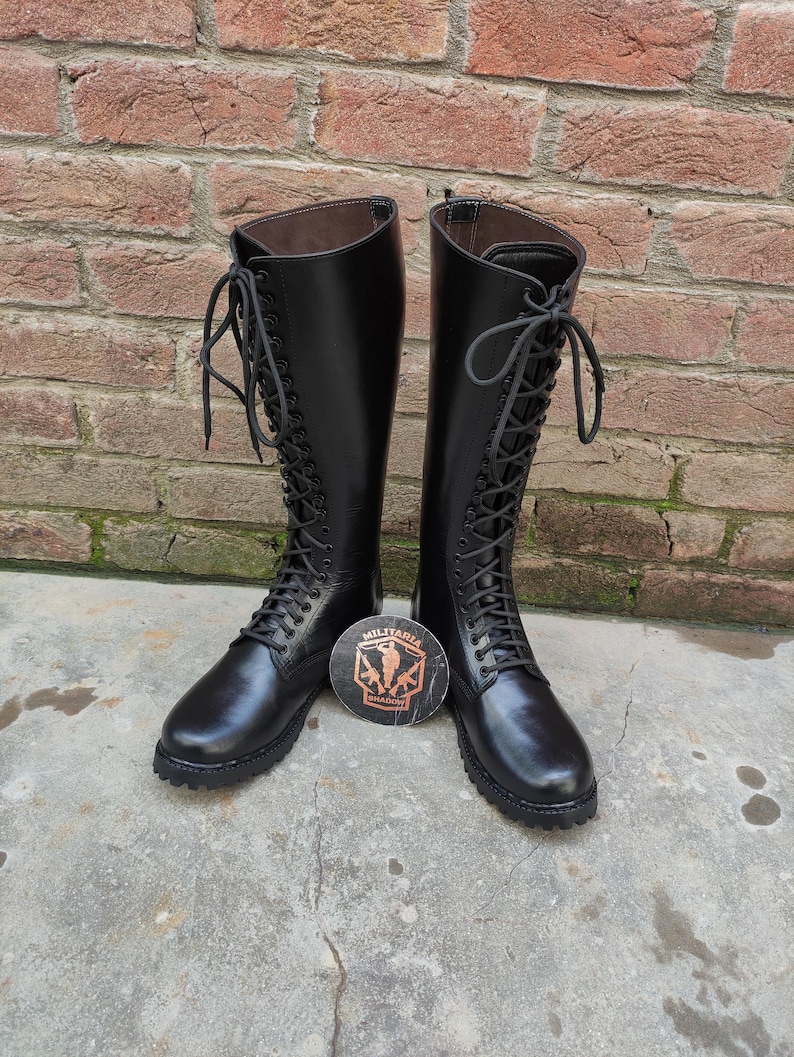 May include: A pair of black leather knee-high boots with lace-up closures. The boots have a rounded toe and a sturdy sole. A round emblem with the text "MILITARIA" is between the boots.