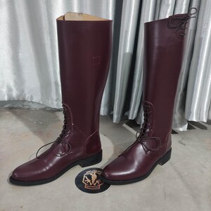 May include: A pair of tall, burgundy leather boots with laces. The boots have a classic design, with a smooth finish and a low heel. The boots are displayed on a light gray surface.
