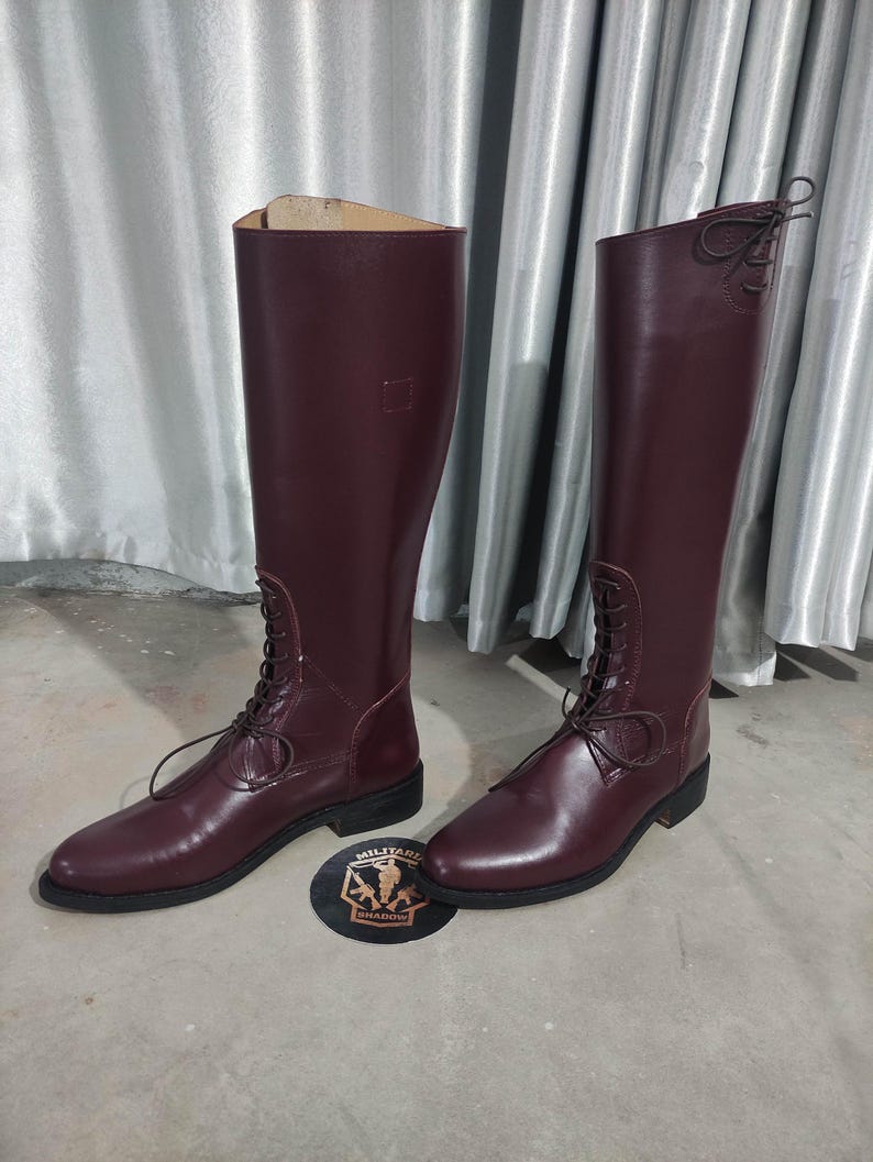 May include: A pair of tall, burgundy leather boots with laces. The boots have a classic design, with a smooth finish and a low heel. The boots are displayed on a light gray surface.
