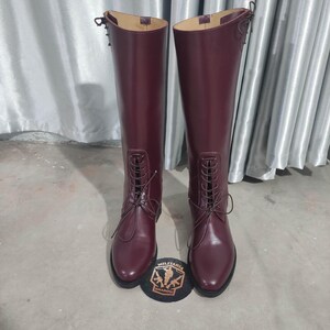 May include: A pair of tall, burgundy leather riding boots with lace-up fronts. The boots have a smooth finish and are displayed with a patch that reads "MILITARIA SHADOW".
