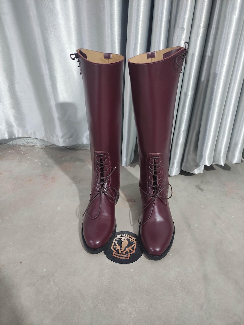 May include: A pair of tall, burgundy leather riding boots with lace-up fronts. The boots have a smooth finish and are displayed with a patch that reads "MILITARIA SHADOW".