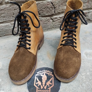 May include: A pair of tan and brown leather boots with black laces. The boots have a suede toe and a leather upper. The boots are displayed on a concrete surface with a circular emblem that reads "Militaria Shadow".