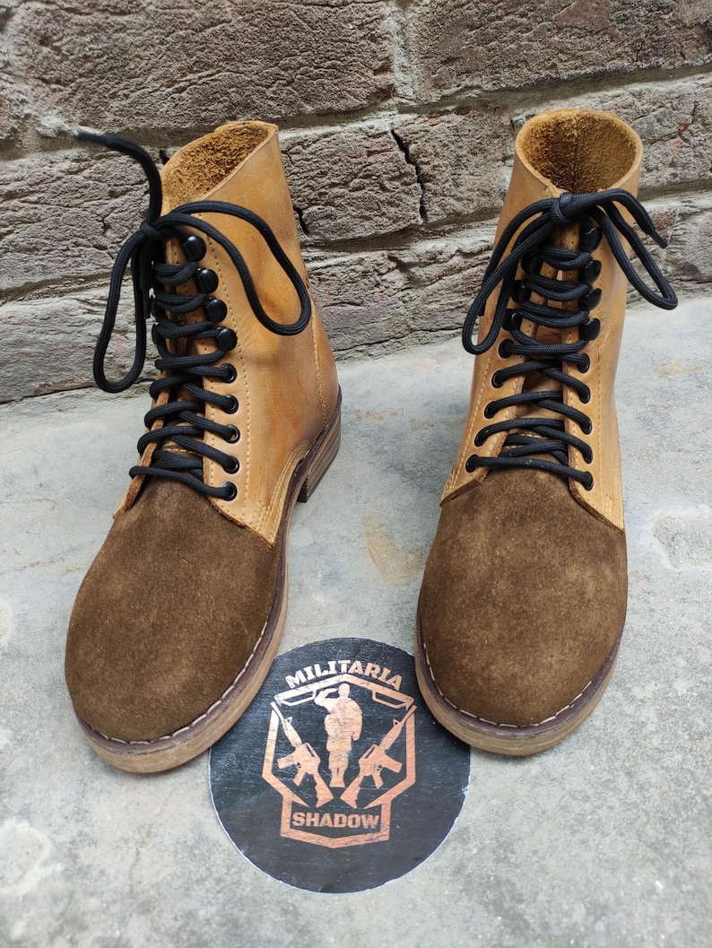 May include: A pair of tan and brown leather boots with black laces. The boots have a suede toe and a leather upper. The boots are displayed on a concrete surface with a circular emblem that reads "Militaria Shadow".