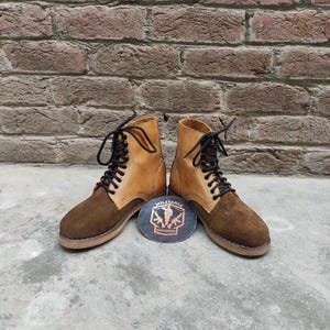 May include: A pair of brown and tan leather boots with black laces. The boots have a suede toe and a leather upper. A round emblem with the text "MILITARIA SHADOW" is between the boots.