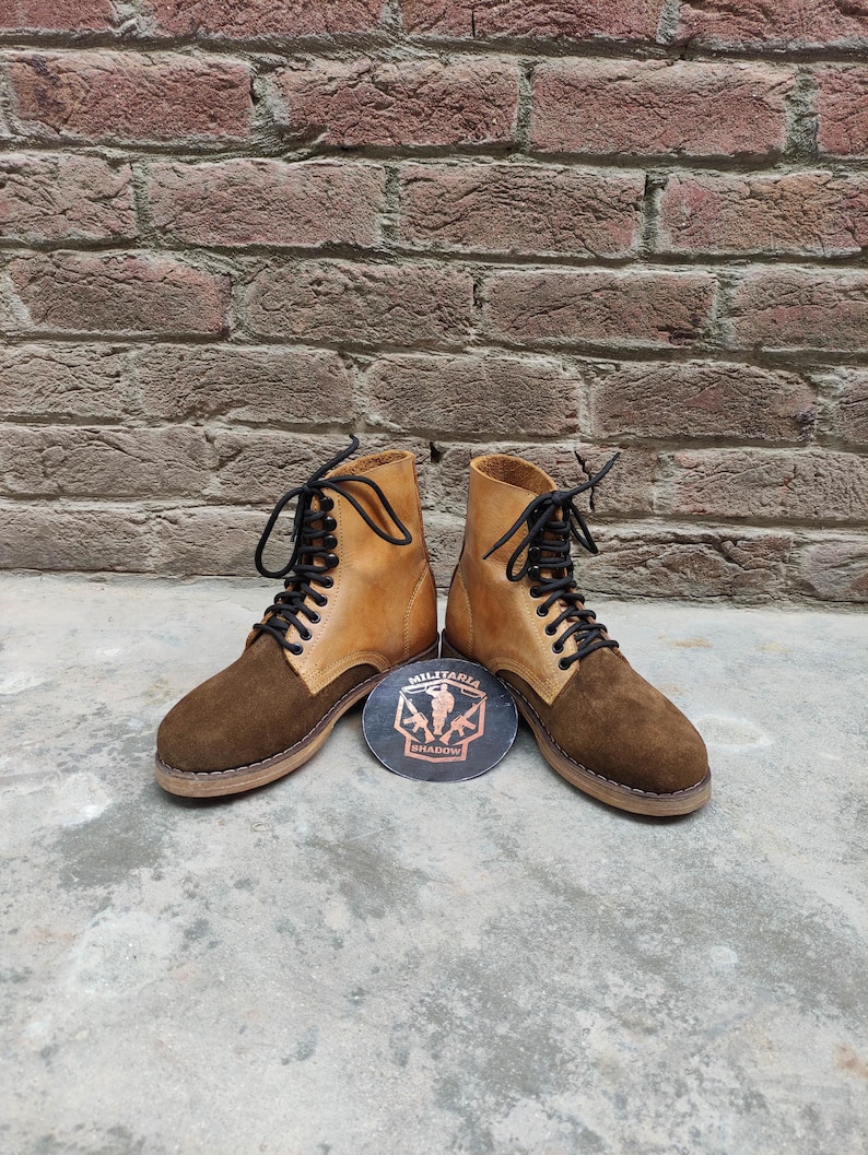 May include: A pair of brown and tan leather boots with black laces. The boots have a suede toe and a leather upper. A round emblem with the text "MILITARIA SHADOW" is between the boots.