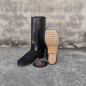 Puede incluir: Botas de montar de cuero negro con empeine de ante y suela tachonada. Las botas son altas, con caña alta, y se exhiben junto a un parche con el texto "MILITARIA SHADOW".