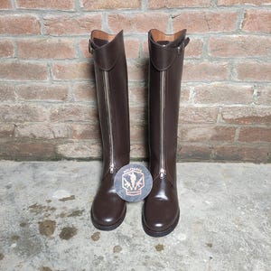 May include: A pair of tall, brown leather riding boots with silver zippers and buckle details. The boots are standing upright on a concrete surface, with a brick wall in the background. The boots have a classic equestrian design.