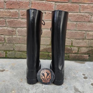 May include: A pair of tall, black leather boots with laces, standing upright against a brick wall. A round emblem with the words "Militaria Shadow" is placed between the boots. The boots have a shiny finish.