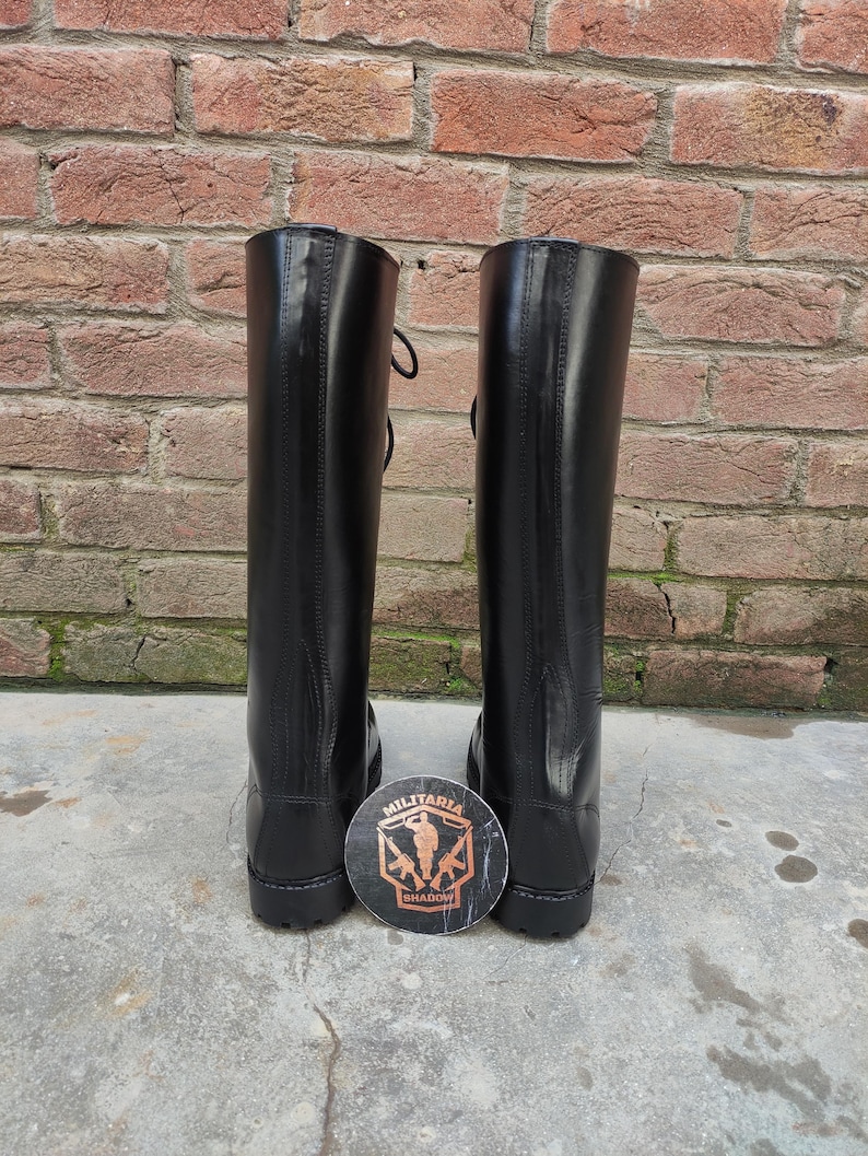 May include: A pair of tall, black leather boots with laces, standing upright against a brick wall. A round emblem with the words "Militaria Shadow" is placed between the boots. The boots have a shiny finish.