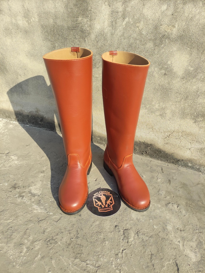 May include: A pair of tall, reddish-brown leather boots. The boots have a smooth finish and a classic design, with a rounded toe and a high shaft. The interior lining is a lighter tan color. A round emblem with the text "MILITARIA SHADOW" is at the base of the boots.