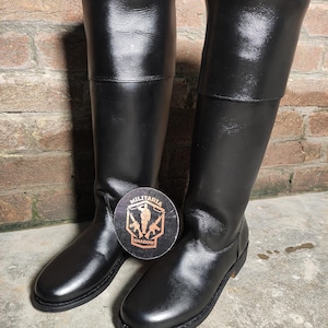 May include: A pair of black leather boots with a tall shaft and a folded-over top. The boots are shiny and appear to be new. A round sticker with the words "MILITARIA SHADOW" and a military-themed graphic is placed between the boots.